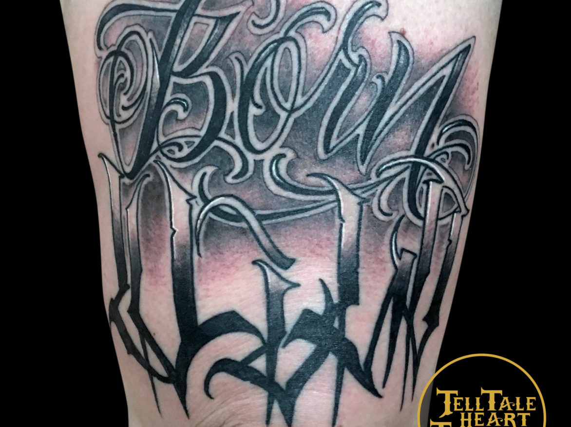 black and grey tattoo of hand drawn script that reads BORN WILD with soft grey background shading on front of thigh