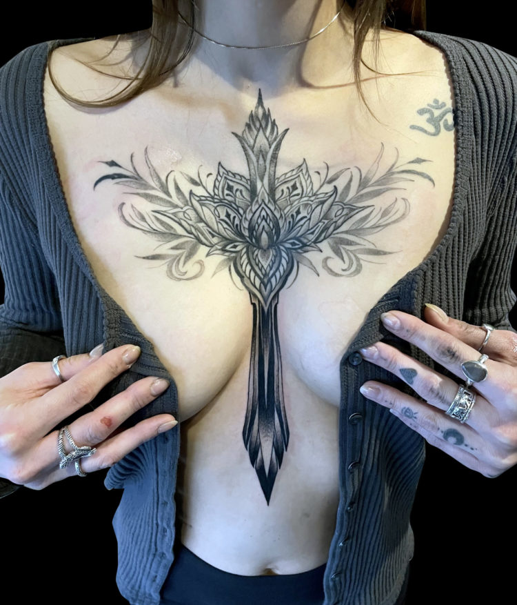 black and grey tattoo of lotus flower with ornamental filigree on chest and sword like shape plunging onto stomach