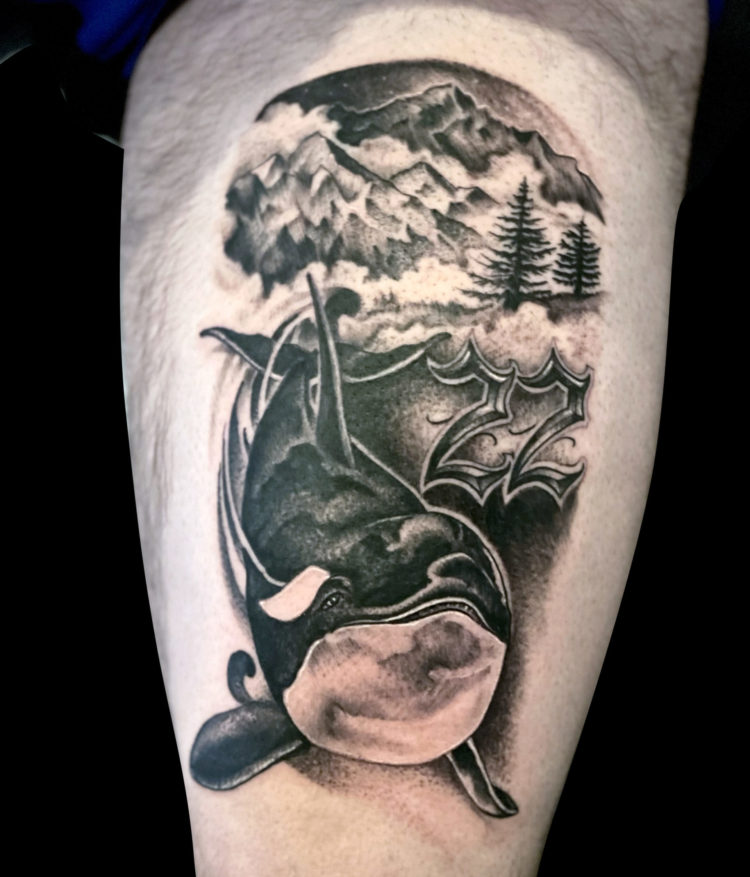 Black and grey tattoo of killer whale orca with mountain range and trees in background and the number 22 in a custom font