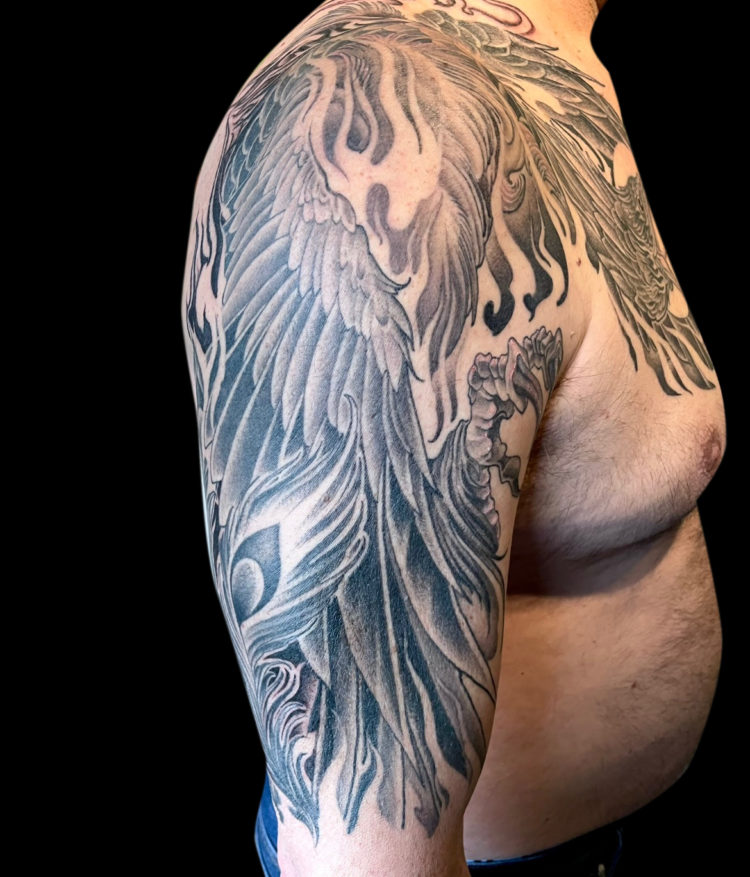 Black and grey tattoo sleeve of phoenix that comes over onto chest with moon in background and negative space flames and lots of feathers