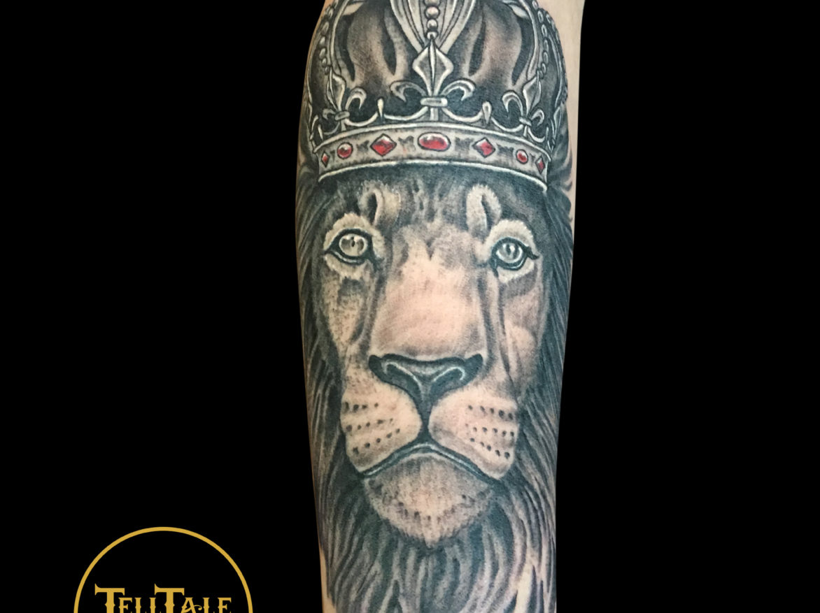 black and grey portrait tattoo of a male lion wearing a king's crown with red jewels on a forearm.
