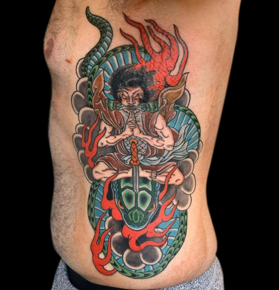 Japanese samurai tattoo seated on green and blue snake head with sword piercing between its eyes. Background is the rest of the coiled snake and flames
