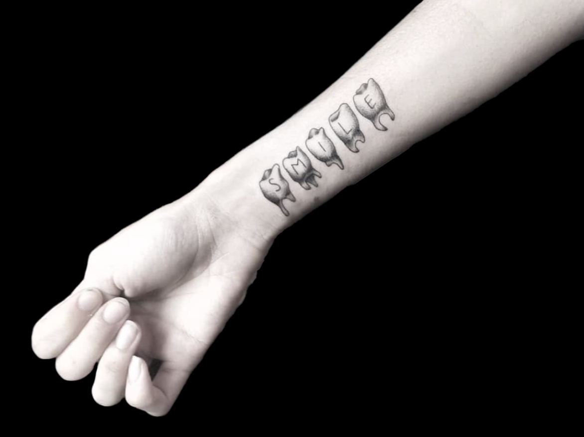 fineline tattoo of five teeth each with a letter that spells out SMILE tattooed on inside wrist
