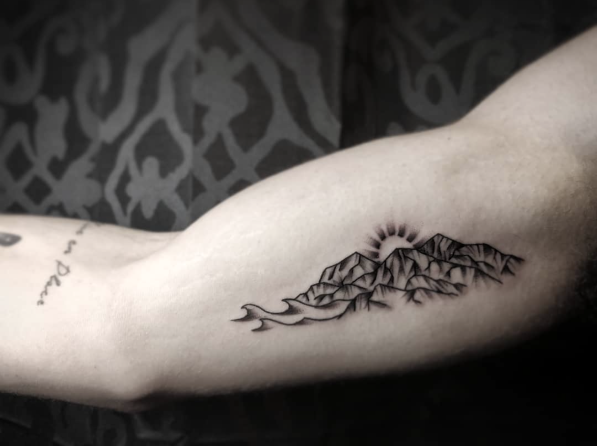 fineline tattoo of mountain range with sun rising behind and simple waves tattooed on inside bicep