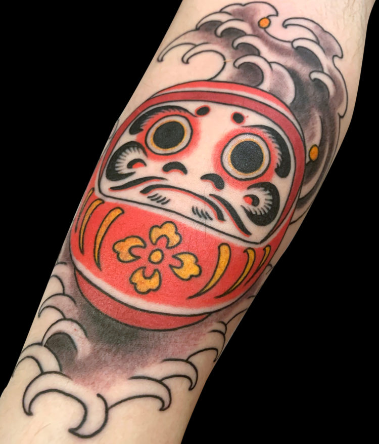 colour tattoo of a round,redJapanese daruma doll wth big eyes and finger waves behind it.