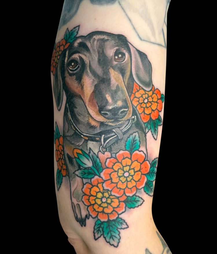 pet memorial colour tattoo of a black and brown dog with flappy ears, sitting wearing a back collar and five orange flowers.