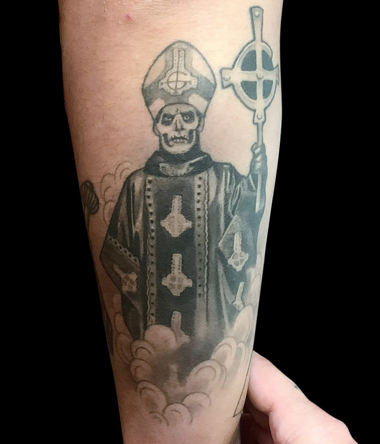 black and grey papa Emeritus Ghost band tattoo featuring Papa character holding staff amidst smoke on forearm