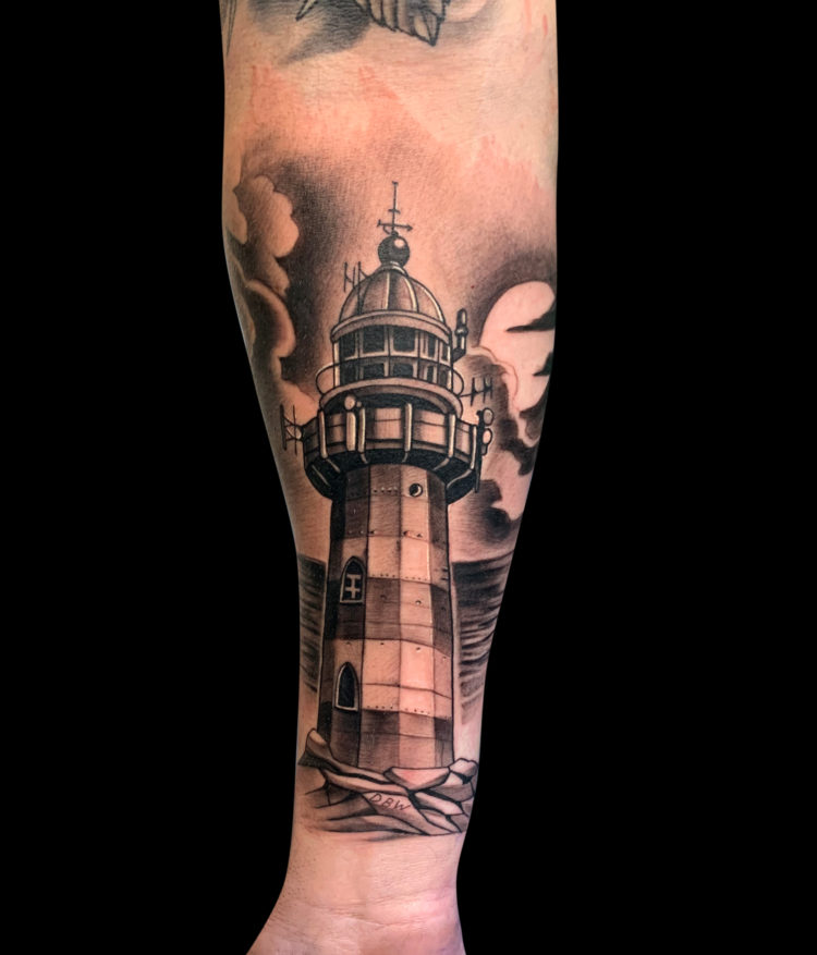 black and grey realism tattoo of lighthouse on rocks with water in background and clouds on forearm