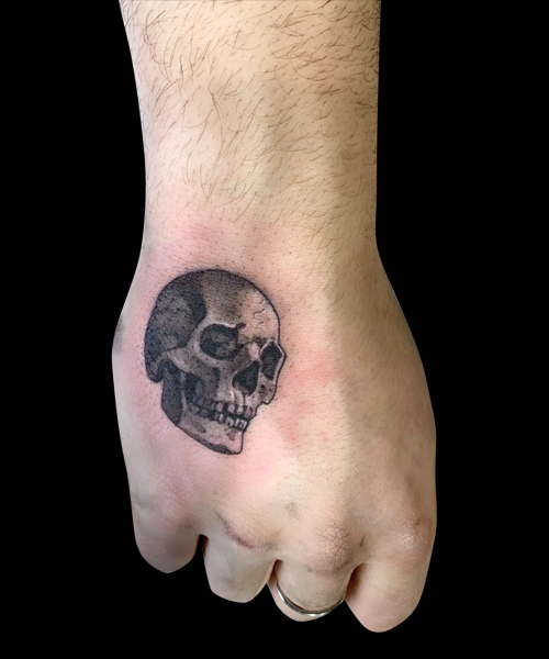 black and grey tattoo of small 3/4 view of skull on hand, taking up less than half of the outside of the top of hand